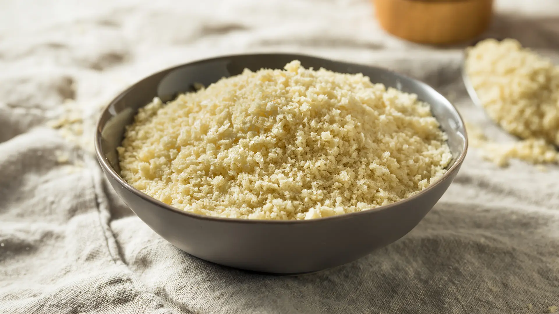 A medium grey bowl filled with Hovis Soft White Medium Sliced breadcrumbs on top of a light grey linen cloth. In the background, a metal spoon with breadcrumbs.