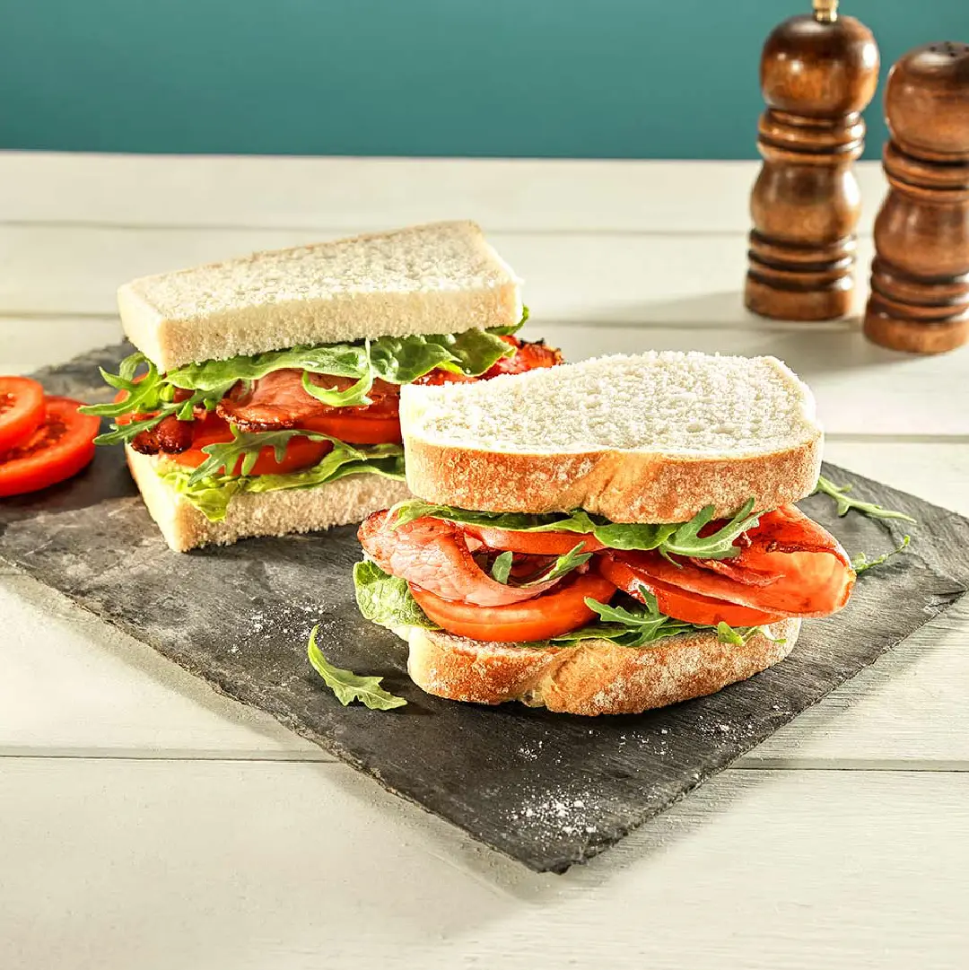 A sandwich cut in half, with crispy bacon, salad leaves and tomato slices between white sliced bread.