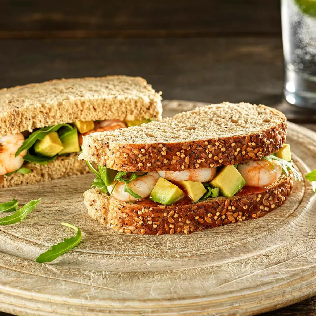 A seeded bread sandwich, sliced in half, filled with prawns, cubed avocado, and rocket leaves.