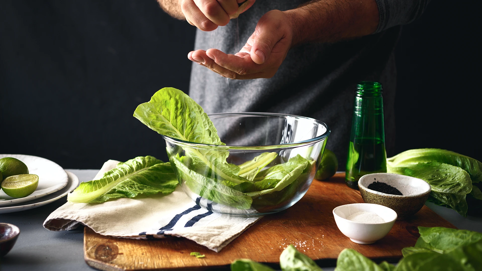 One hand squeezing a lemon over a glass bowl of green salad leaves, while the other hand forms a shell to catch any seeds.