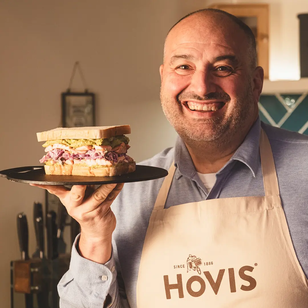 A man in a Hovis apron holds a plate with a sandwich filled with mashed avocado, salmon, and coleslaw.