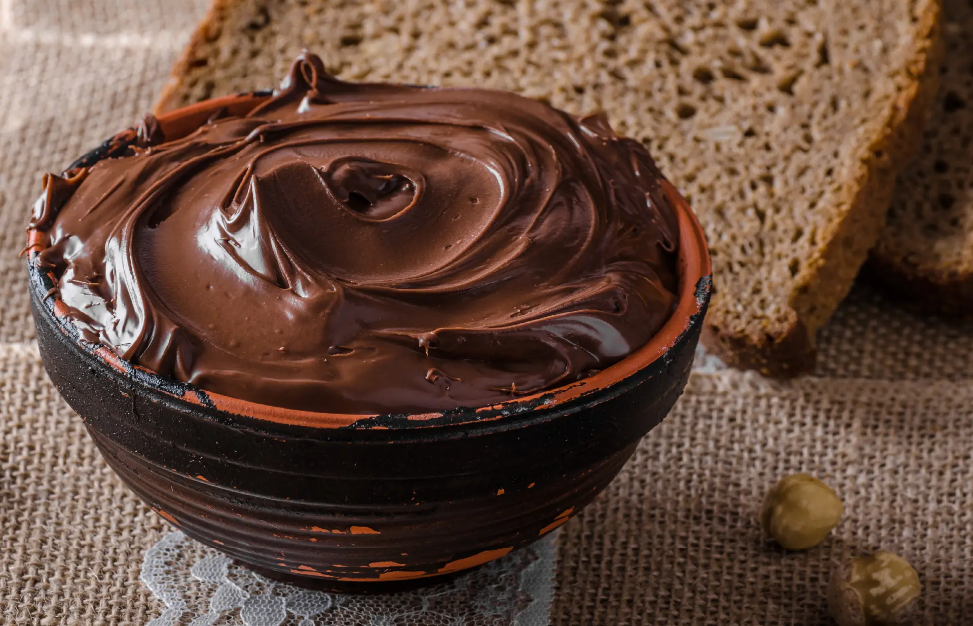 A bowl filled with smooth brown chocolate spread, with slices of brown bread in the background.