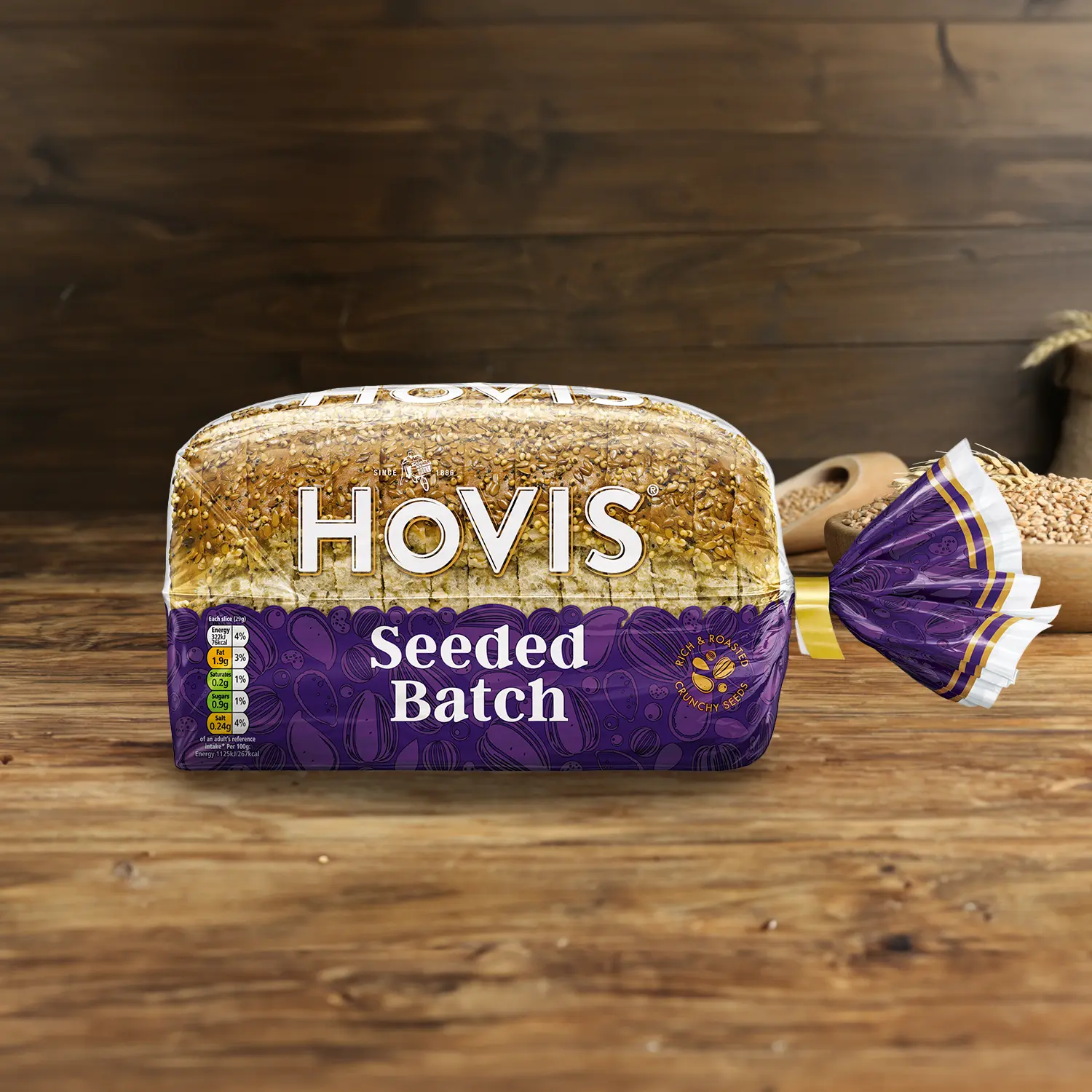 Hovis Seeded Batch loaf on a wooden table, with a bowl of wheat grains, a wooden scoop, and dried wheat in the background.