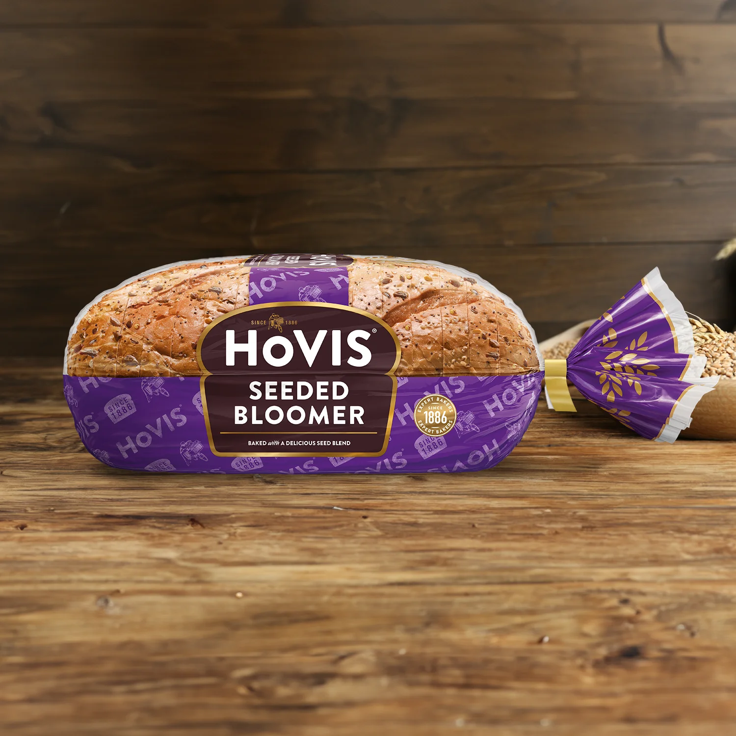 Hovis Seeded Bloomer on a wooden table, with a bowl of wheat grains, a wooden scoop, and dried wheat in the background.