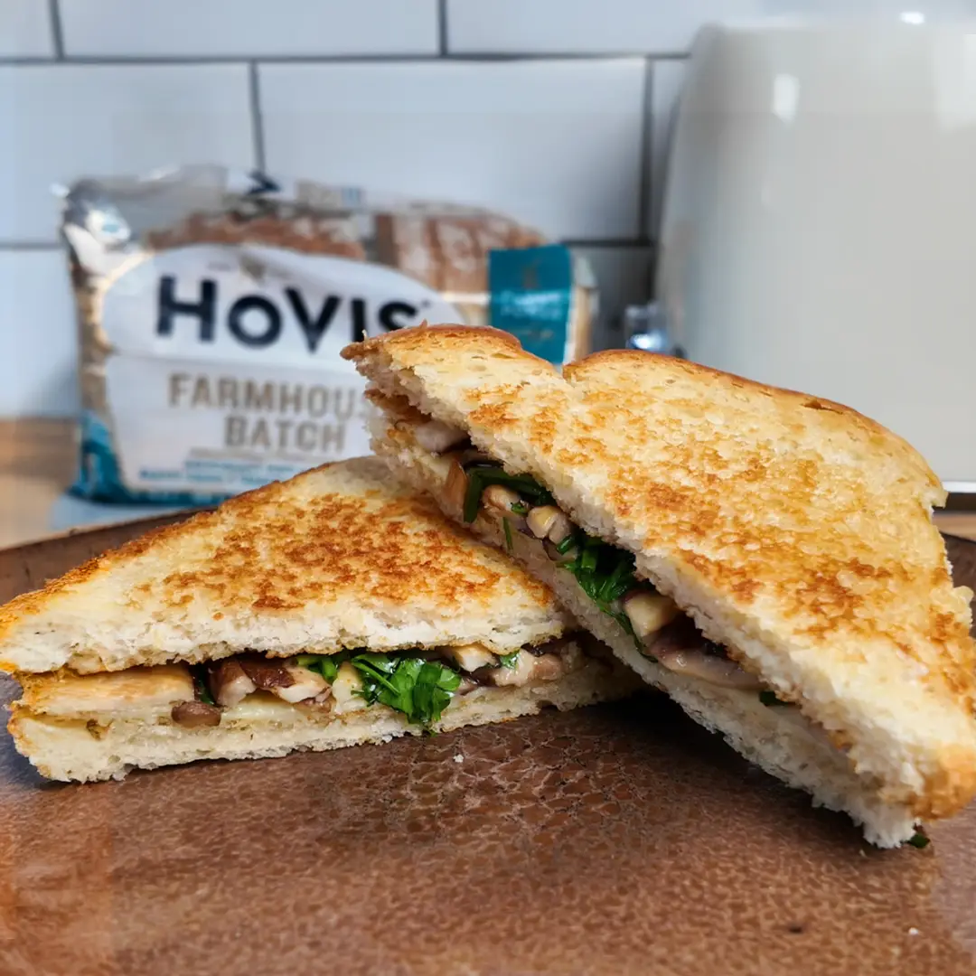 A golden-brown toasted sandwich cut in half, revealing a filling of sautéed mushrooms, greens, and melted cheese. In the background, a Hovis Farmhouse Batch bread pack is visible on a kitchen counter.