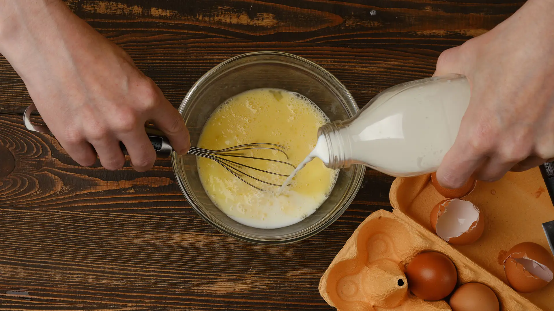 Eggs and milk being whisked together in a glass bowl on a wooden countertop.