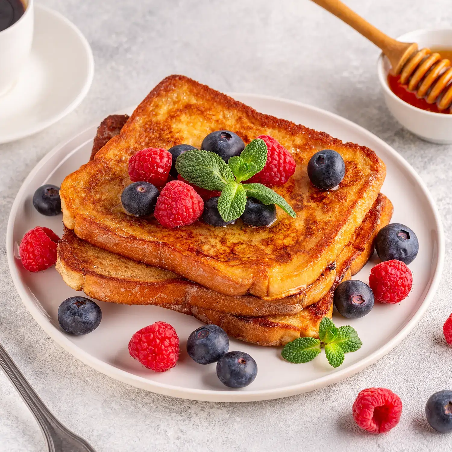 A plate on a white wooden chopping board, with three golden Hovis Soft White Bread French toasts stacked. Some blueberries, raspberries, honey and a mint leaf on top.