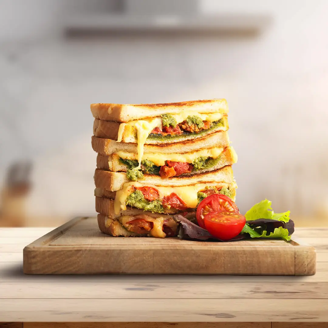 Four halves of pesto and cheese toasties stacked on a wooden chopping board, with molten cheese and pesto slightly visible between the slices. A halved tomato and some salad leaves are placed to the right.