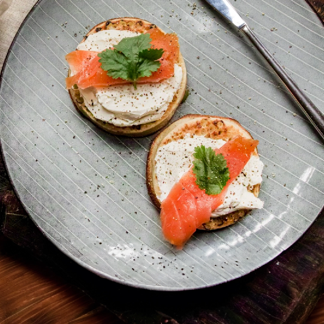 Top view of two slightly toasted crumpets topped with cream cheese, a thin slice of smoked salmon, a parsley leaf, and ground pepper.