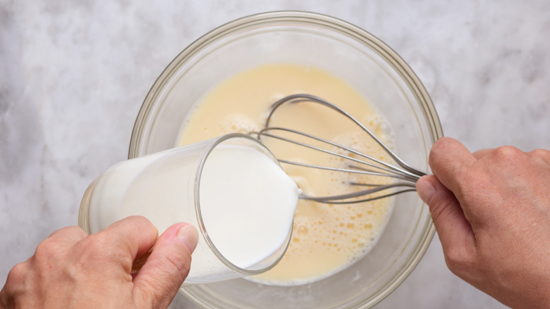 One hand holding a small glass jug, pouring milk into a glass bowl with beaten eggs, while the other hand uses a whisk to mix the ingredients.