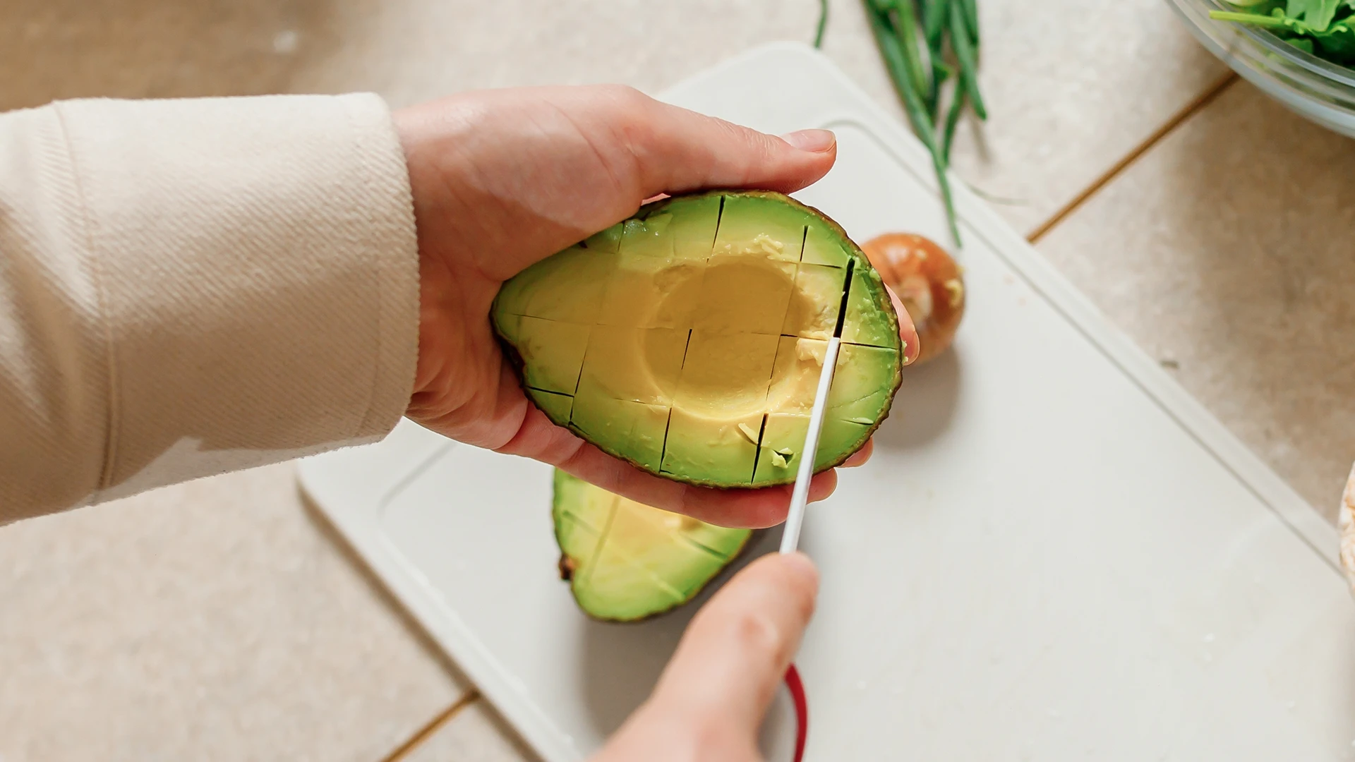 A hand holds half an avocado while the other uses a knife to slice it into cubes.