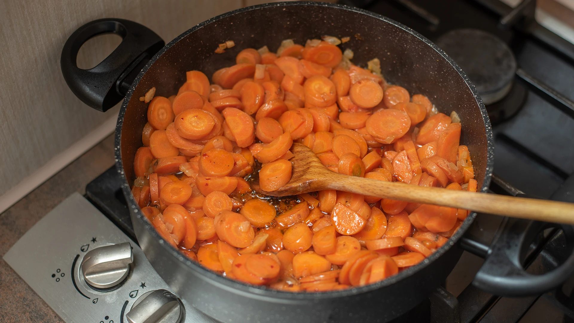 A large pot filled with round carrot slices, chopped onions, and liquid stock, with a wooden spoon inside.