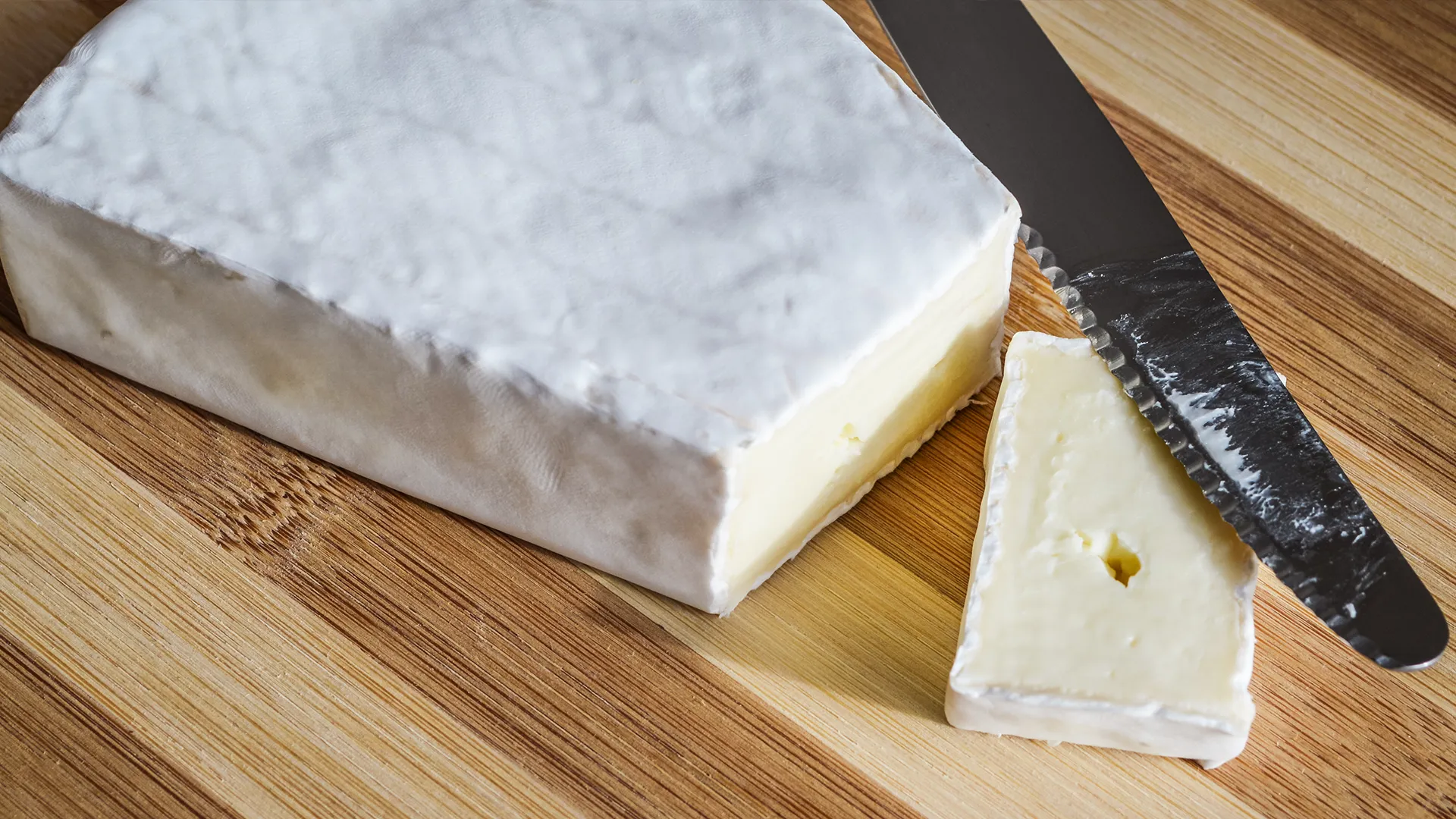 A thin slice of Brie, with a metal knife resting on top, sits on a wooden chopping board, with the remaining Brie alongside it.