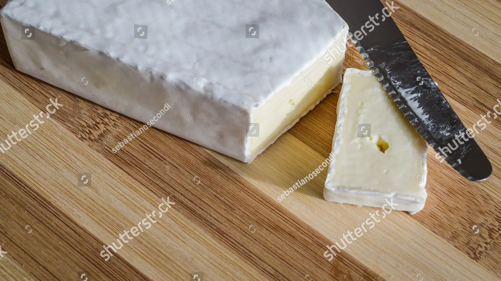 A thin slice of Brie, with a metal knife resting on top, sits on a wooden chopping board, with the remaining Brie alongside it.