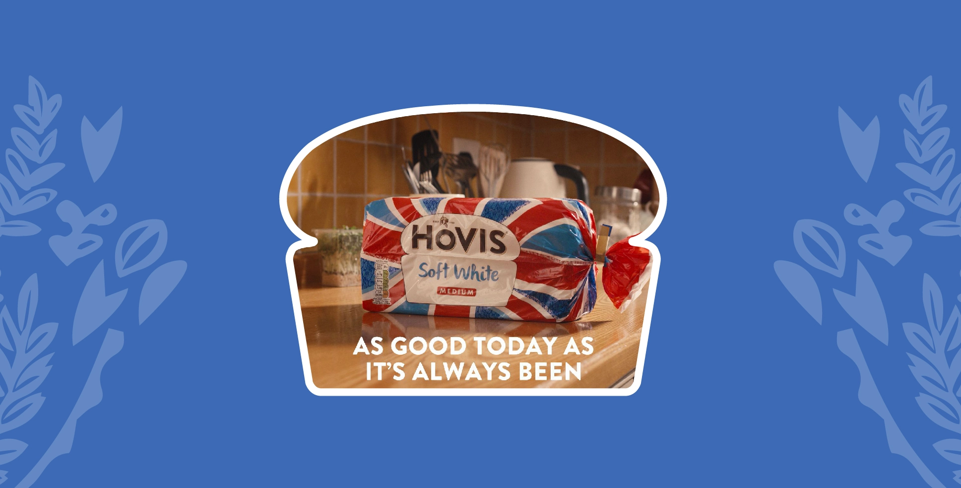 On a blue background with light blue wheats on either side, a Hovis Soft White loaf sits in the middle of the image on top of a kitchen counter, cropped into a loaf shape with a white border.