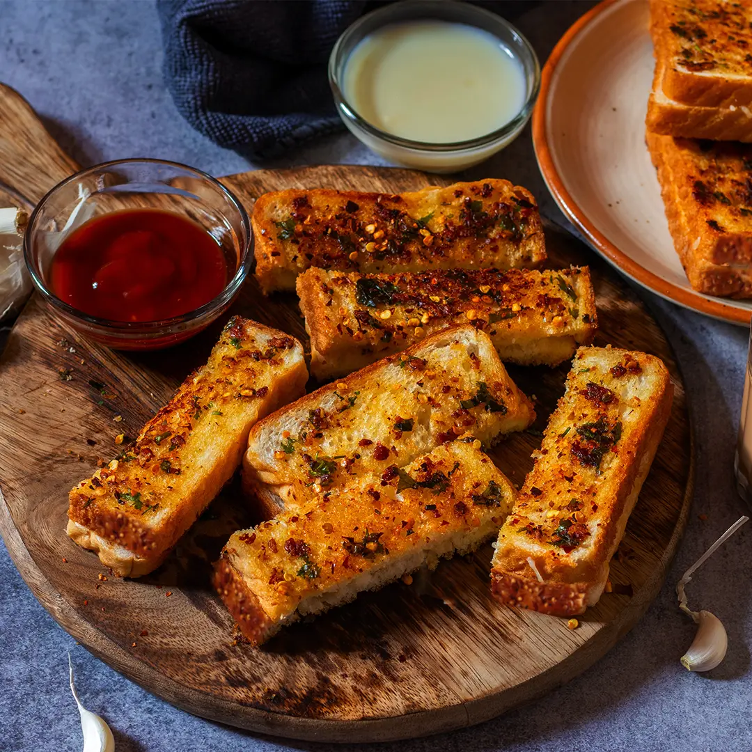 Hovis Seed Sensations Original bread, sliced in half and placed on a wooden chopping board. The slices are golden brown, topped with melted cheese and a sprinkle of thinly sliced herbs. On the side, a small bowl of ketchup and a few garlic cloves.