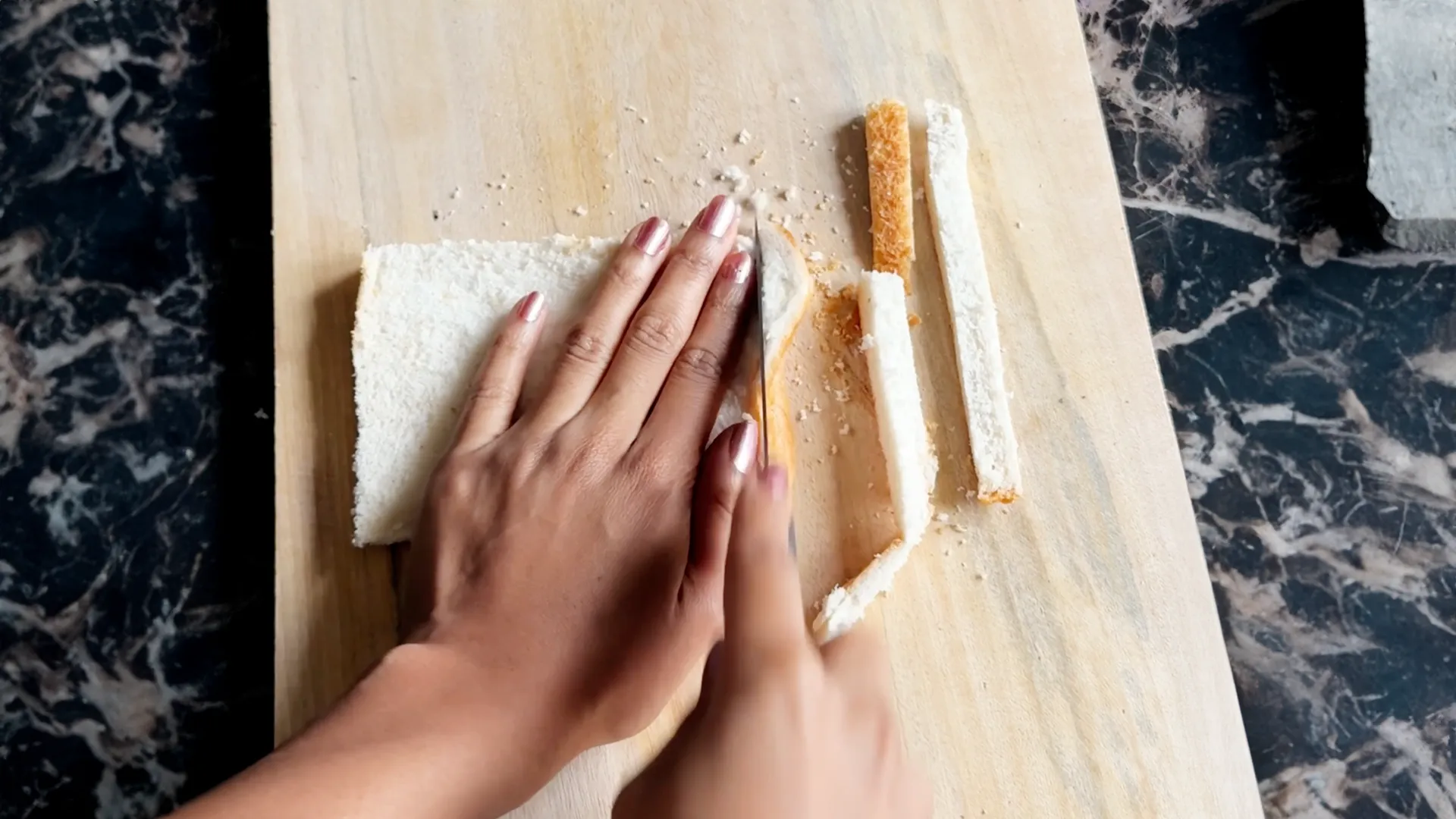 On a wooden chopping board, someone is cutting the crusts off a slice of bread on a dark marble worktop.