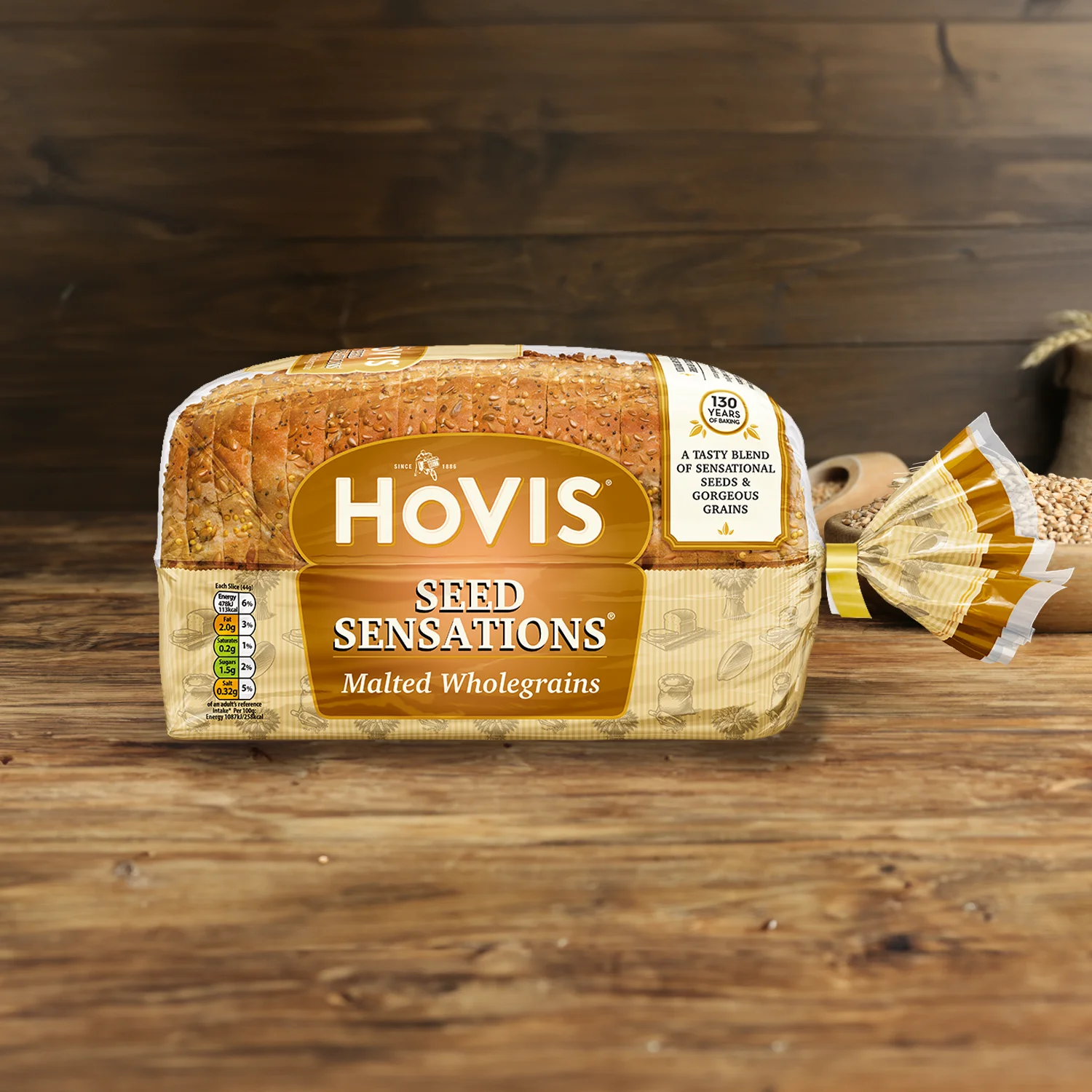 Hovis Seed Sensations Malted Wholegrain on a wooden table, with a bowl of wheat grains, a wooden scoop, and dried wheat in the background.