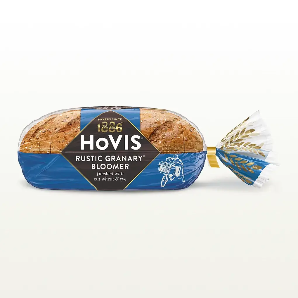 Hovis Rustic Granary Bloomer bread on a white backdrop with a subtle shadow. The packaging is half transparent and half dark blue, decorated with golden wheats.