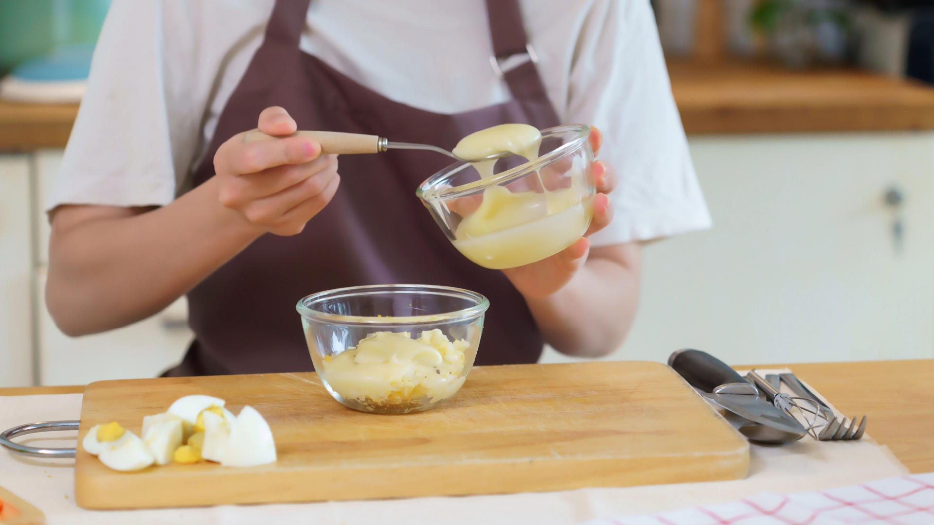 A hand adding a spoonful of mayonnaise to a glass bowl filled with eggs and salad cream.