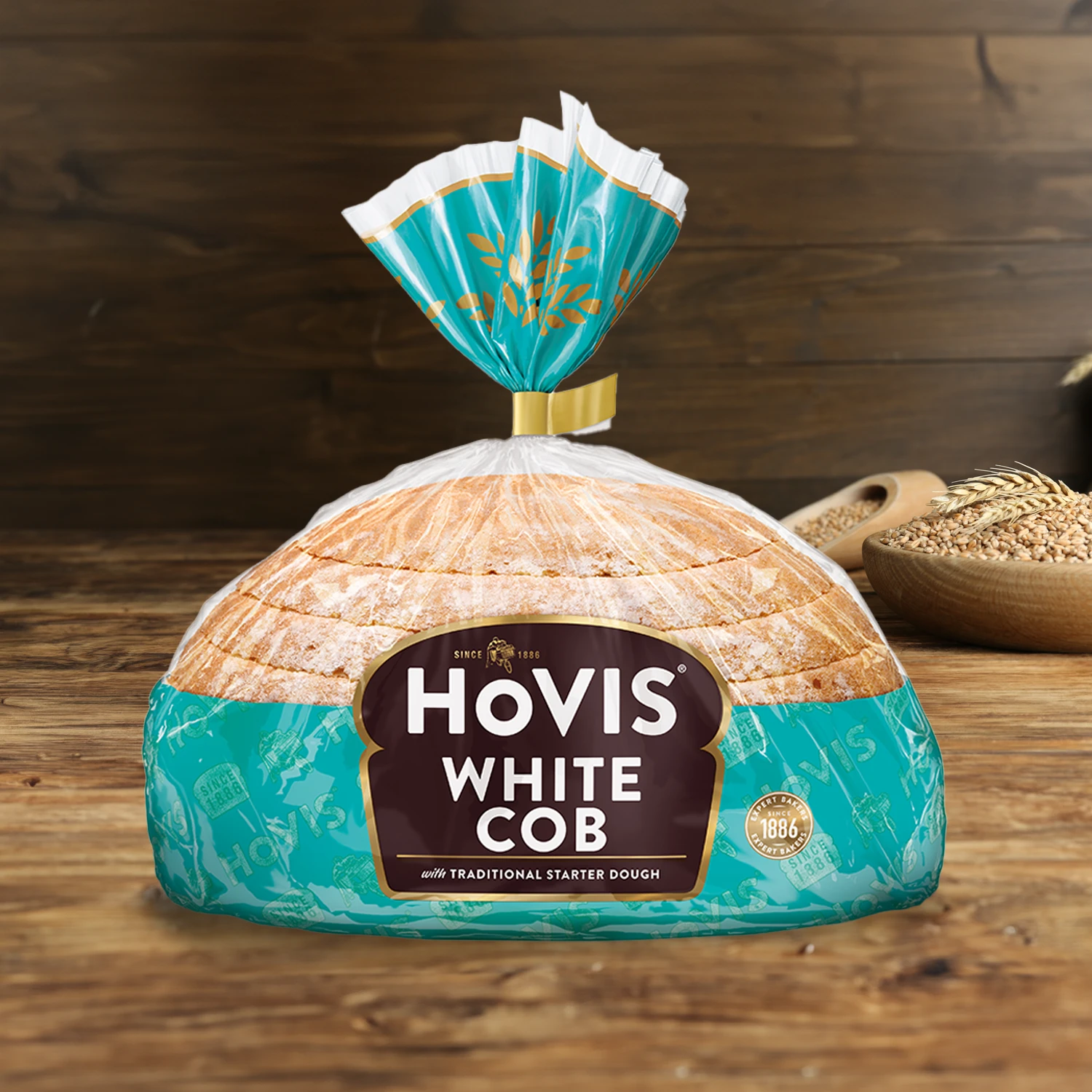 Hovis White Half Cob on a wooden table, with a bowl of wheat grains, a wooden scoop, and dried wheat in the background.