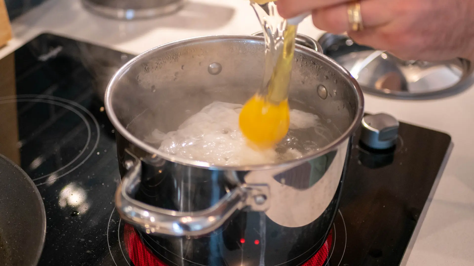 A medium pot of boiling water on a hob, with hands cracking an egg into the pot.