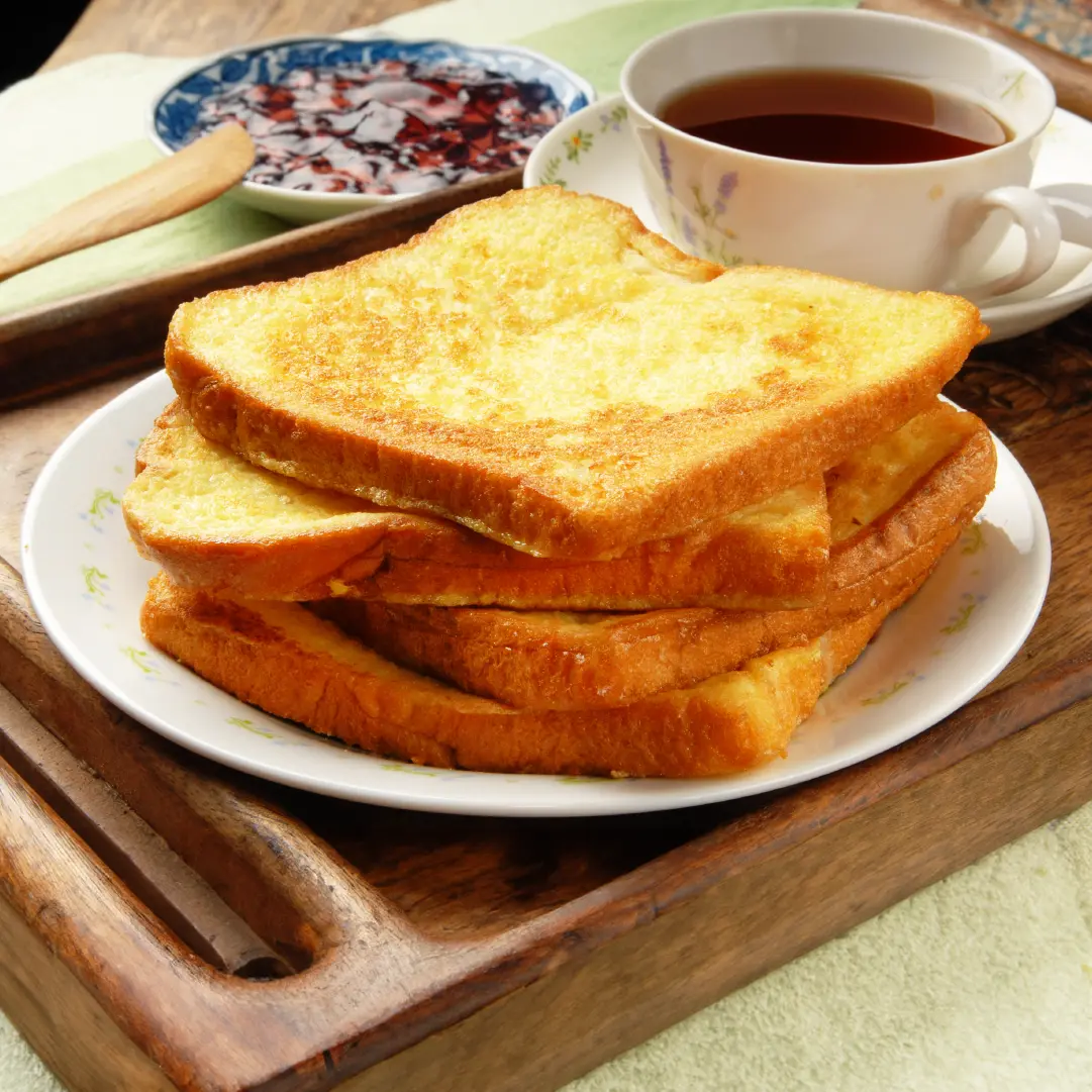 Four slices of toasted white bread stacked on a plate, with a cup of tea and a small plate of jam on the side.