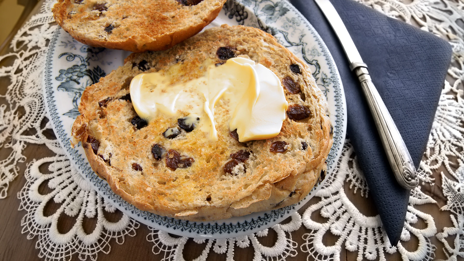 A buttered teacake on a decorative plate with a knife next to it.