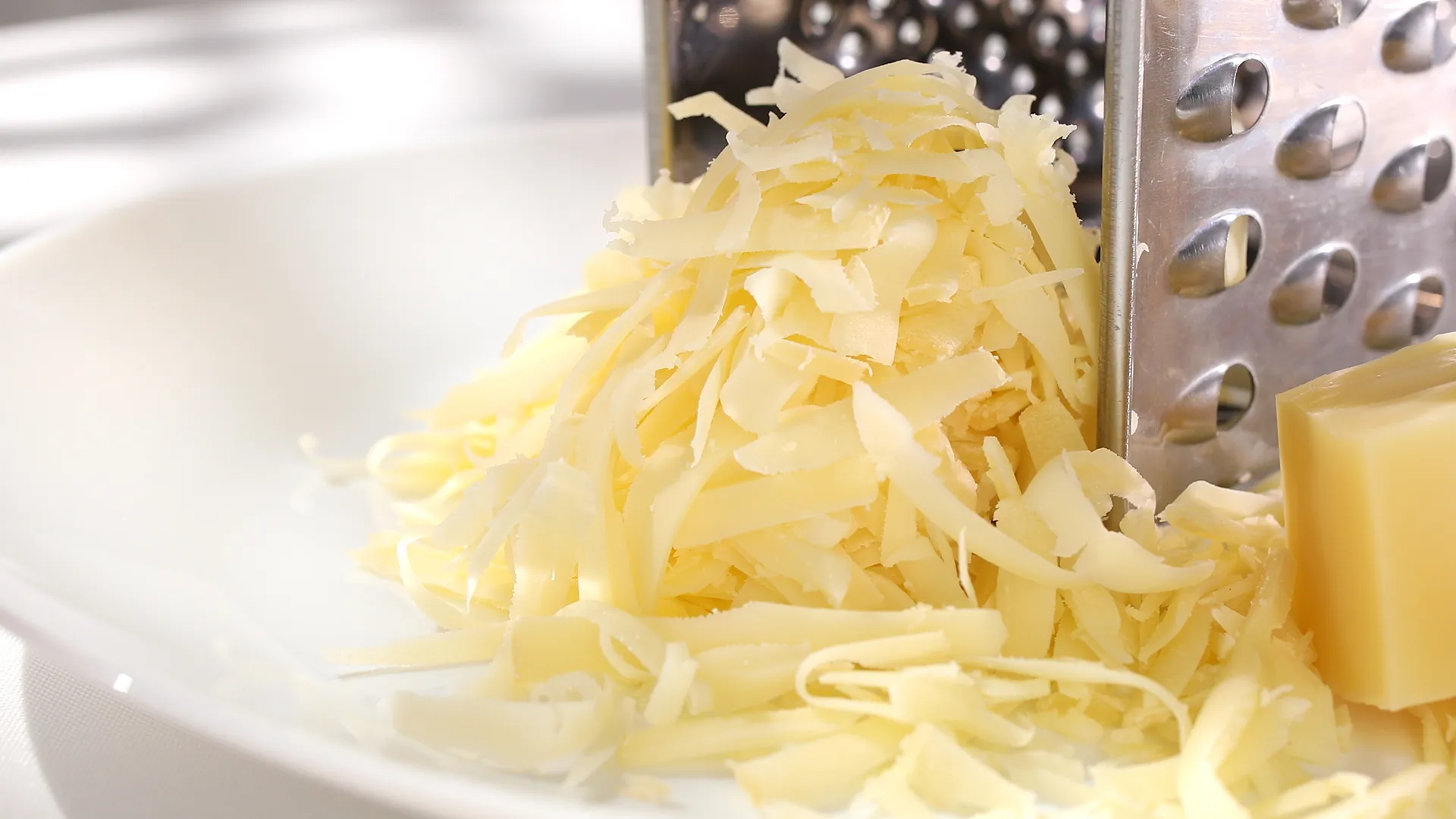 One hand holds a block of cheese, while the other has a metal cheese grater with a wooden handle. Grated cheese falls on a wooden chopping board.