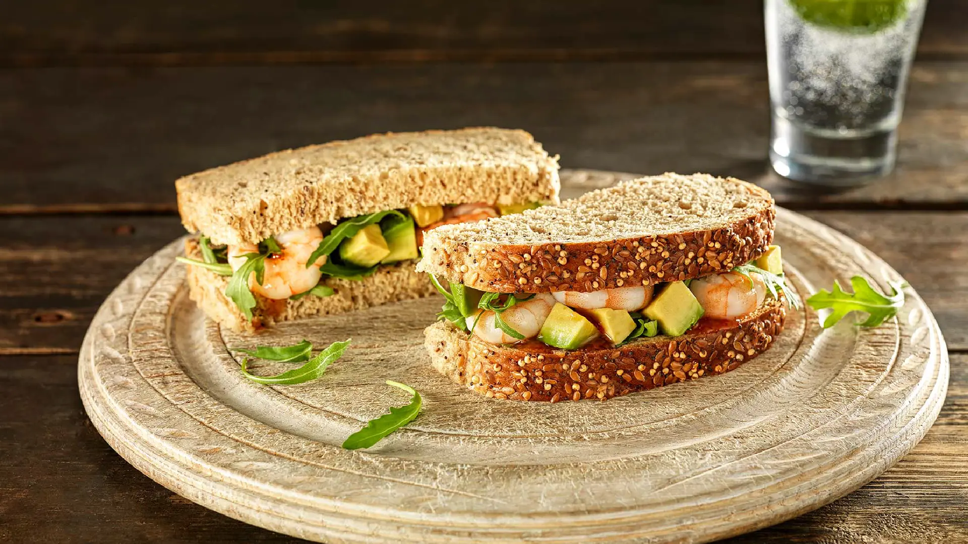 A seeded bread sandwich, sliced in half, filled with prawns, cubed avocado, and rocket leaves.