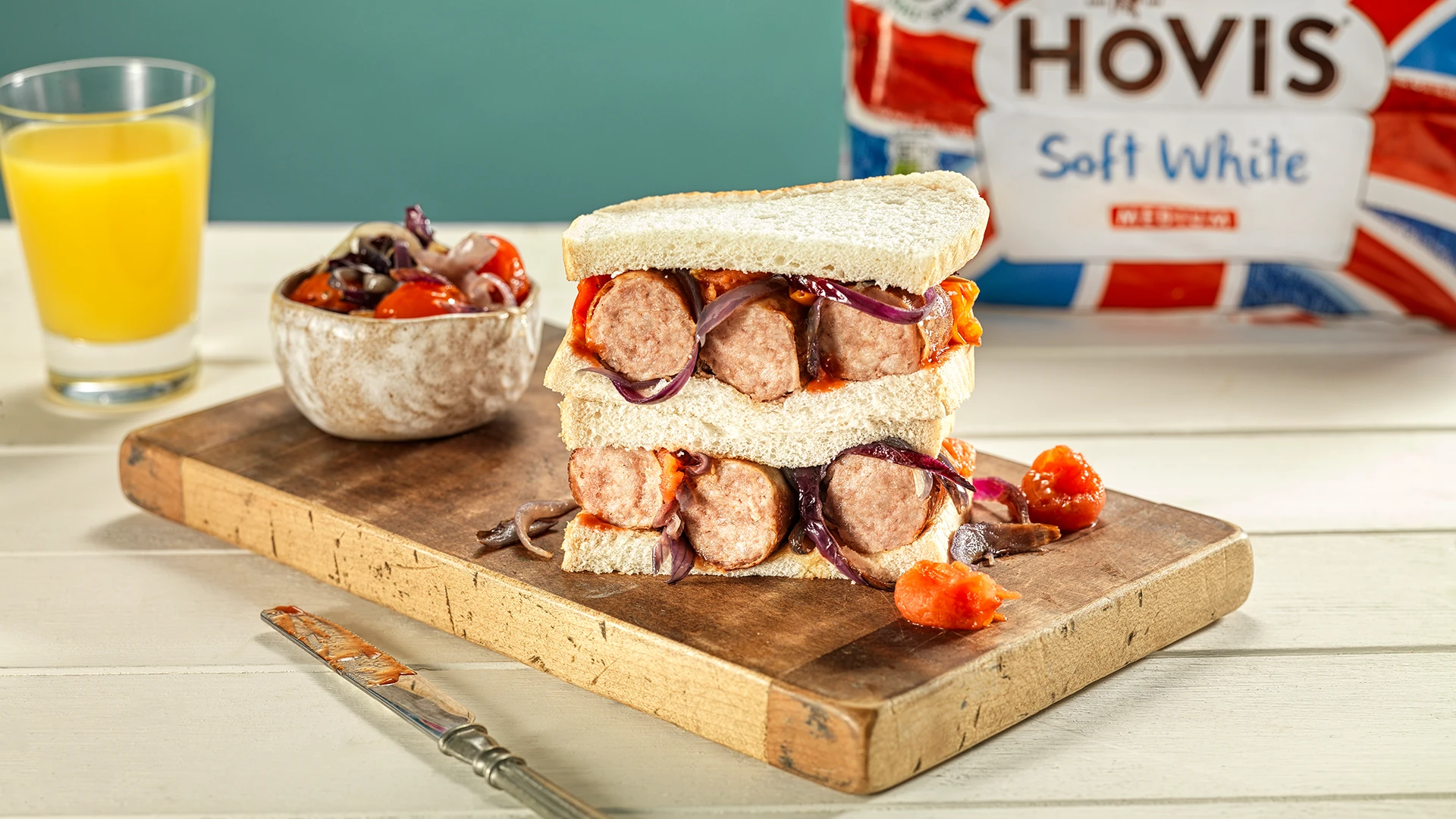 Two halves of a sandwich made with Hovis Soft White Medium Sliced bread, stacked and filled with red onions, sausage, and roasted tomatoes, on a wooden chopping board.