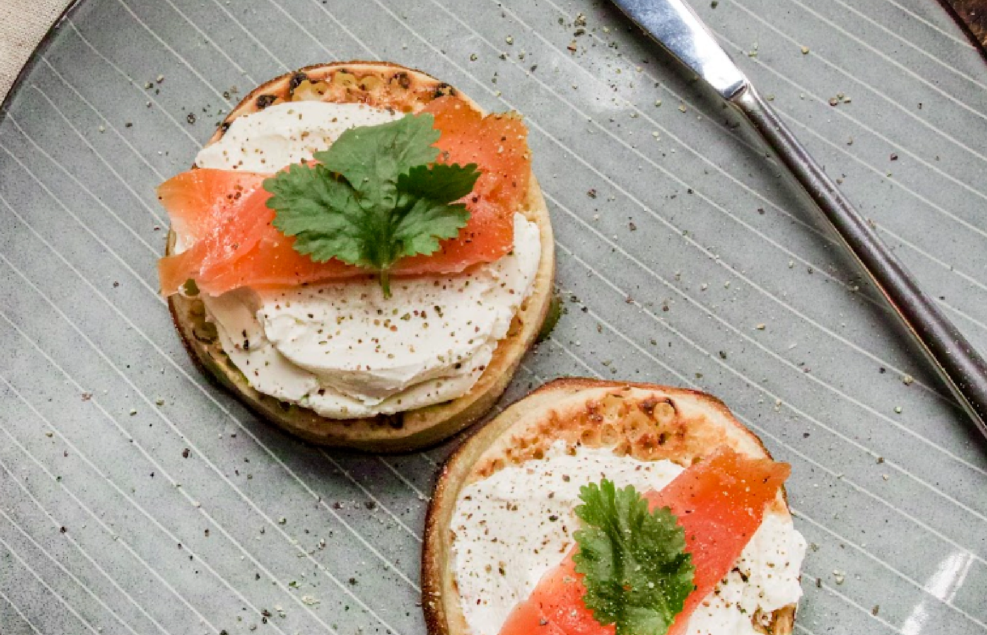 Top view of two slightly toasted crumpets topped with cream cheese, a thin slice of smoked salmon, a parsley leaf, and ground pepper.