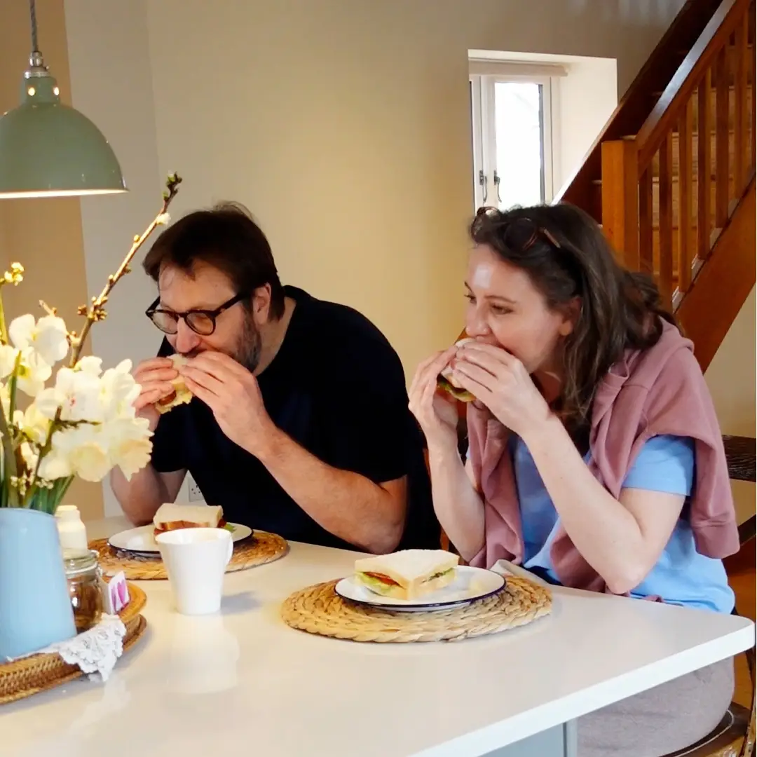 A couple sits by a kitchen counter and eats a delicious sandwich.