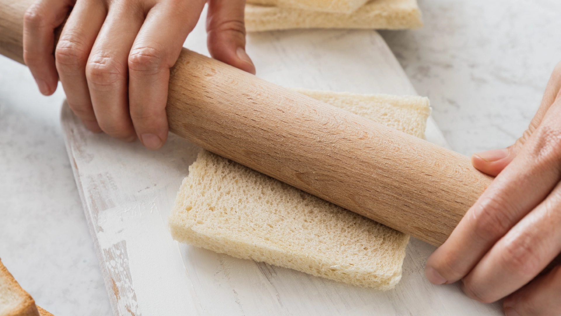 Two hands using a wooden rolling pin to flatten a crustless slice of white bread.