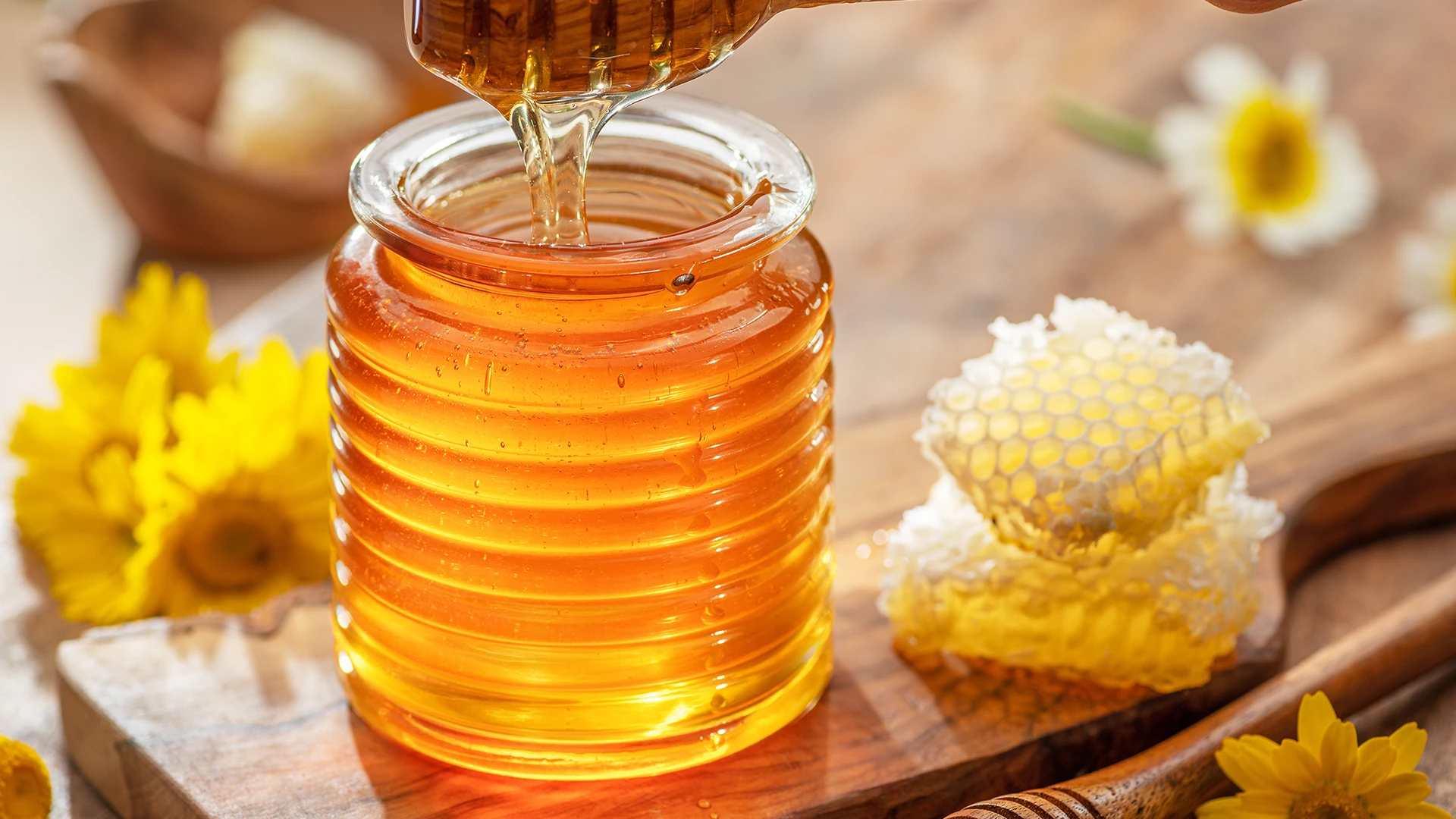 A hand lifting a honey drizzler filled with honey from a glass honey pot.