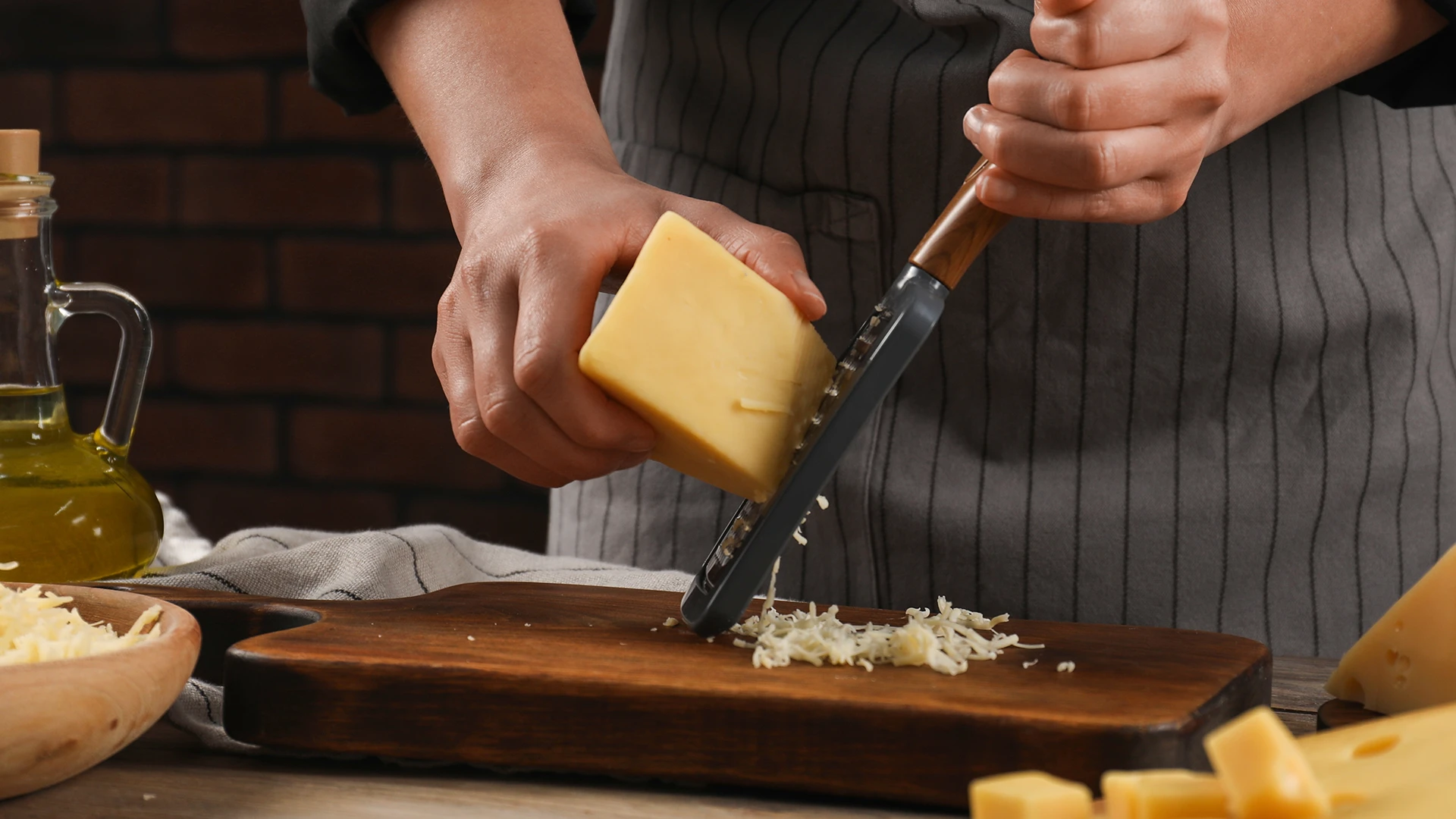 A hand holds a grater at an angle over a wooden chopping board while the other hand grates a piece of yellow cheese.