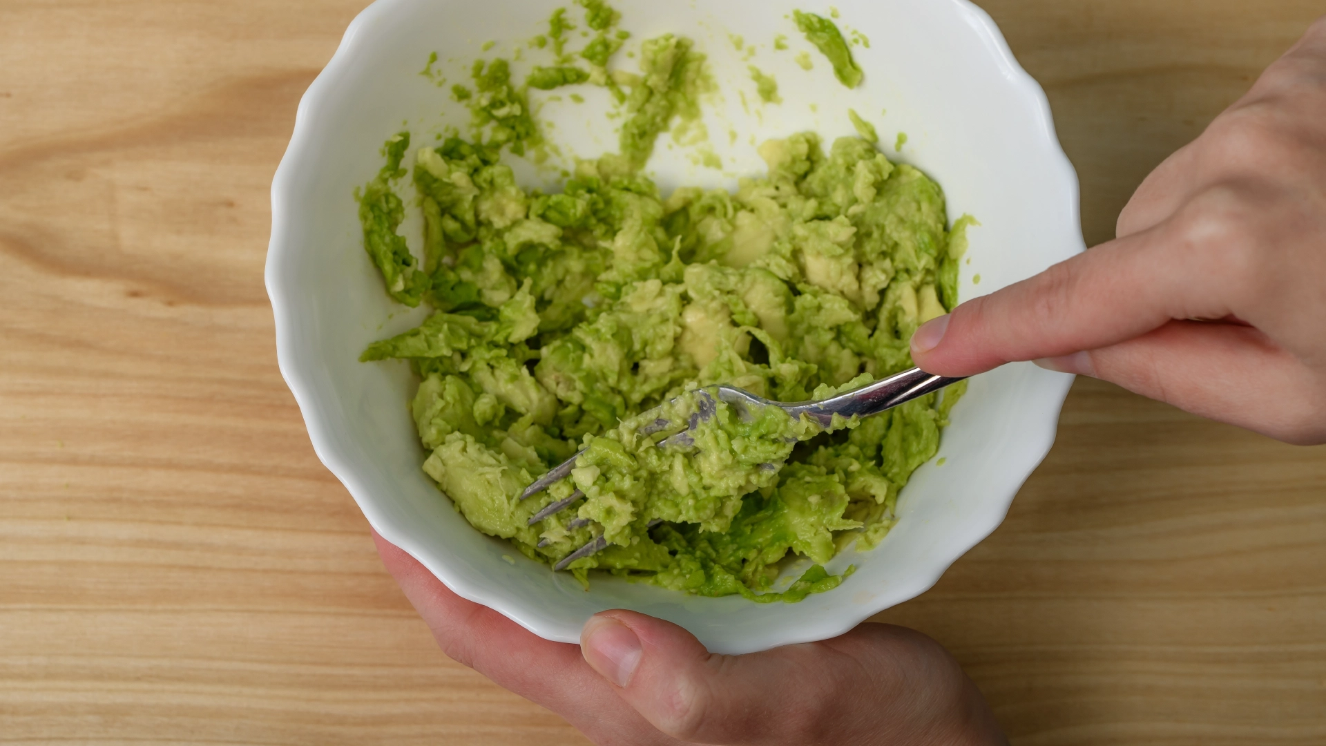 One hand holding a bowl of avocado while the other mashed them with a metal fork.