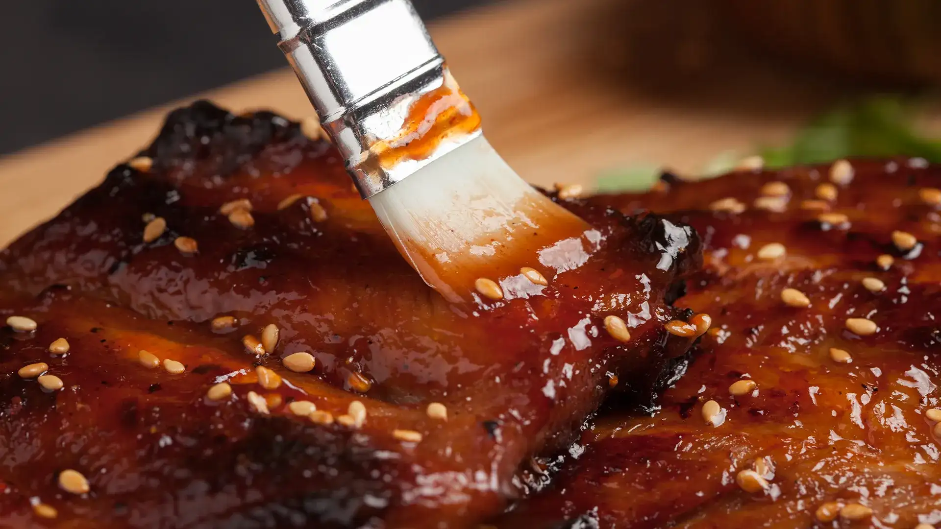 Grilled pork being brushed with a glaze and sesame seeds.