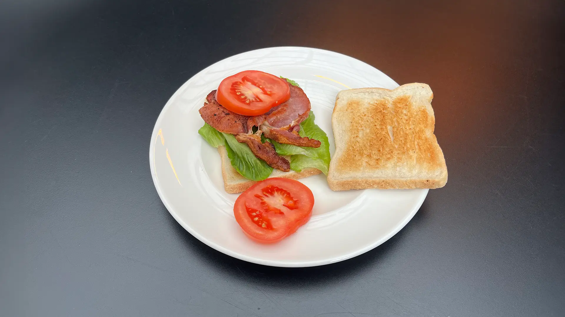 A hand holds a slice of bacon while the other slices it in half with a knife on a wooden chopping board. In the background, there are stacked slices of bread, lettuce leaves, and tomato slices.