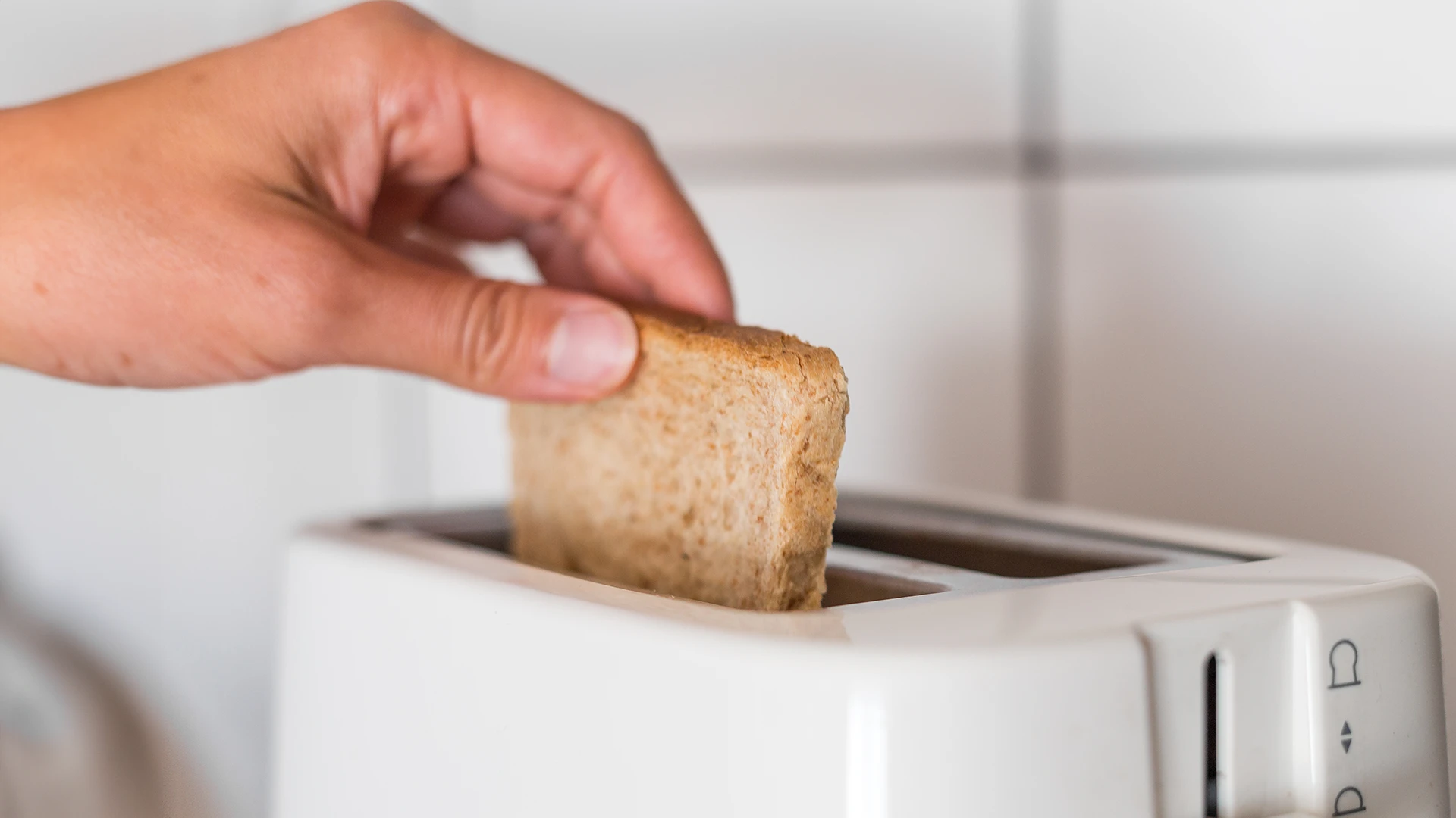 A hand places a slice of brown bread into a toaster.