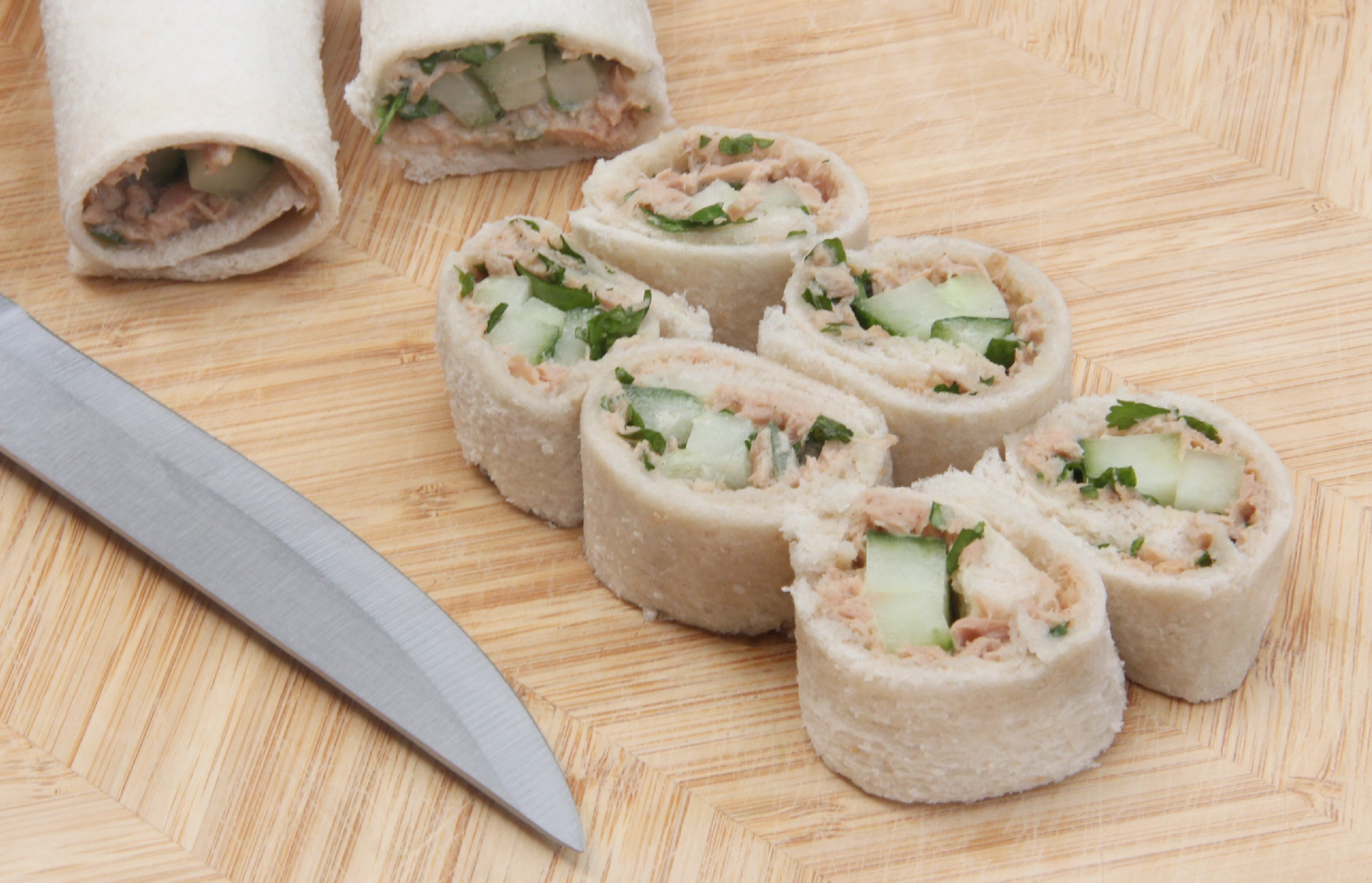 Thin bread wrapped around sliced cucumber and tuna mayo, cut into six bite-sized pieces resembling sushi, with two uncut rolls in the background and a sharp knife.