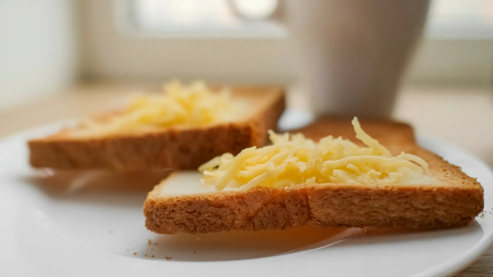 A closed up image of two slices of toasted bread topped with some shredded Gruyère cheese on a white plate. On a wooden kitchen counter.