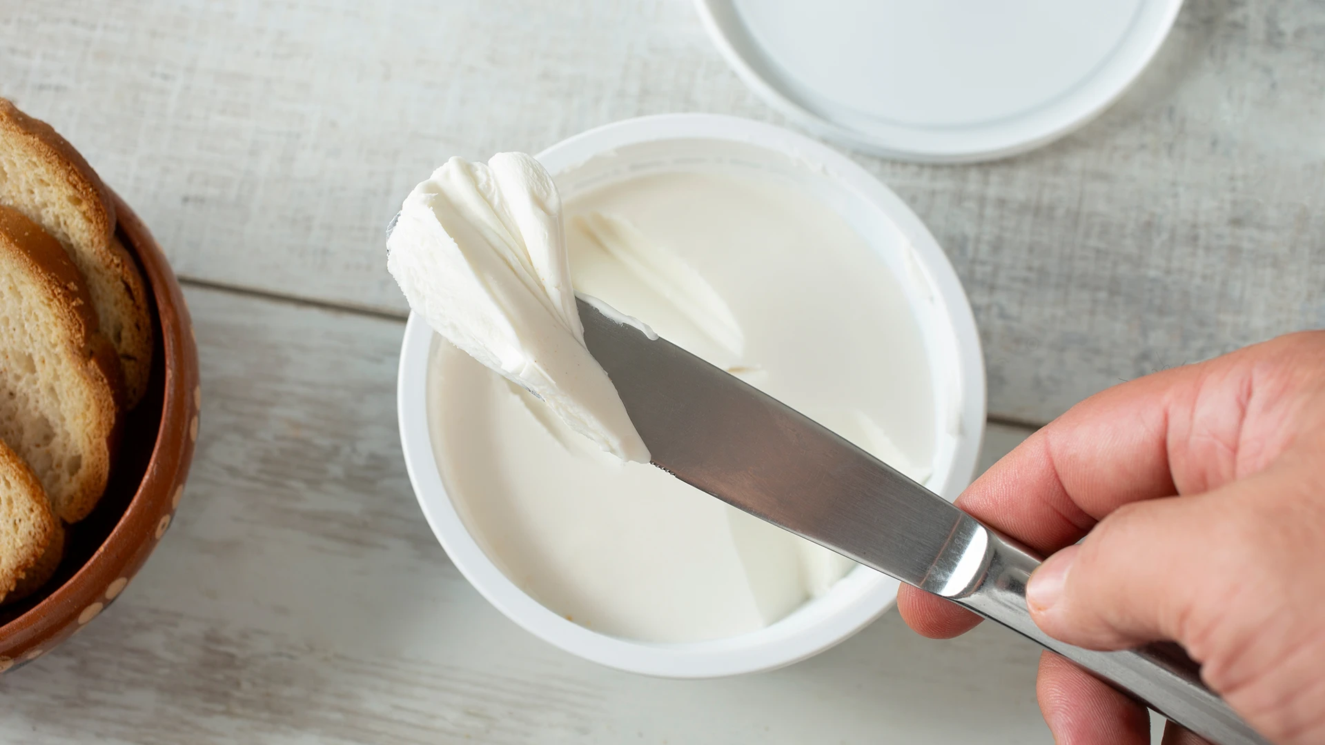 A hand holding a knife to scoop white cream cheese from a pot.